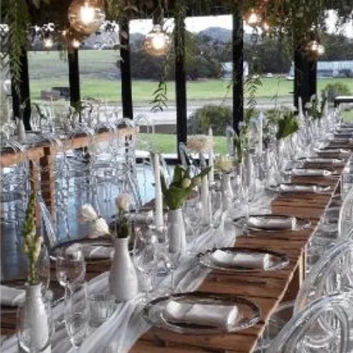 Long wooden tables set with glassware, white bud vases and hanging greenery